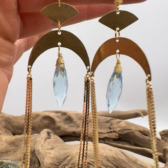 Gold statement earrings with a faceted aqua crystal, a celestial moon charm, and flowing golden tassels. Bold, lightweight, and designed for movement and shine.