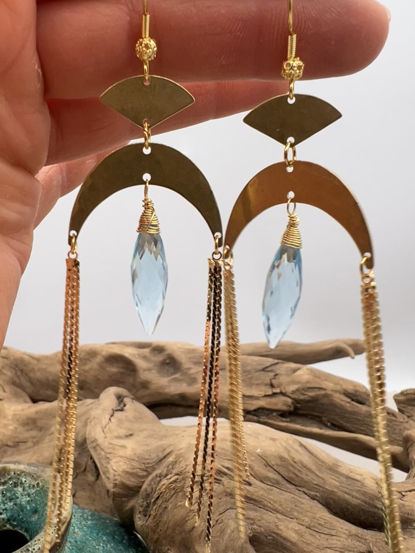 Gold statement earrings with a faceted aqua crystal, a celestial moon charm, and flowing golden tassels. Bold, lightweight, and designed for movement and shine.