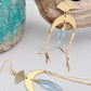 Gold statement earrings with a faceted aqua crystal, a celestial moon charm, and flowing golden tassels. Bold, lightweight, and designed for movement and shine.