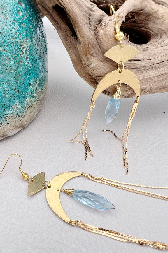 Gold statement earrings with a faceted aqua crystal, a celestial moon charm, and flowing golden tassels. Bold, lightweight, and designed for movement and shine.