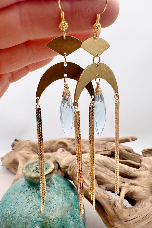 Gold statement earrings with a faceted aqua crystal, a celestial moon charm, and flowing golden tassels. Bold, lightweight, and designed for movement and shine.