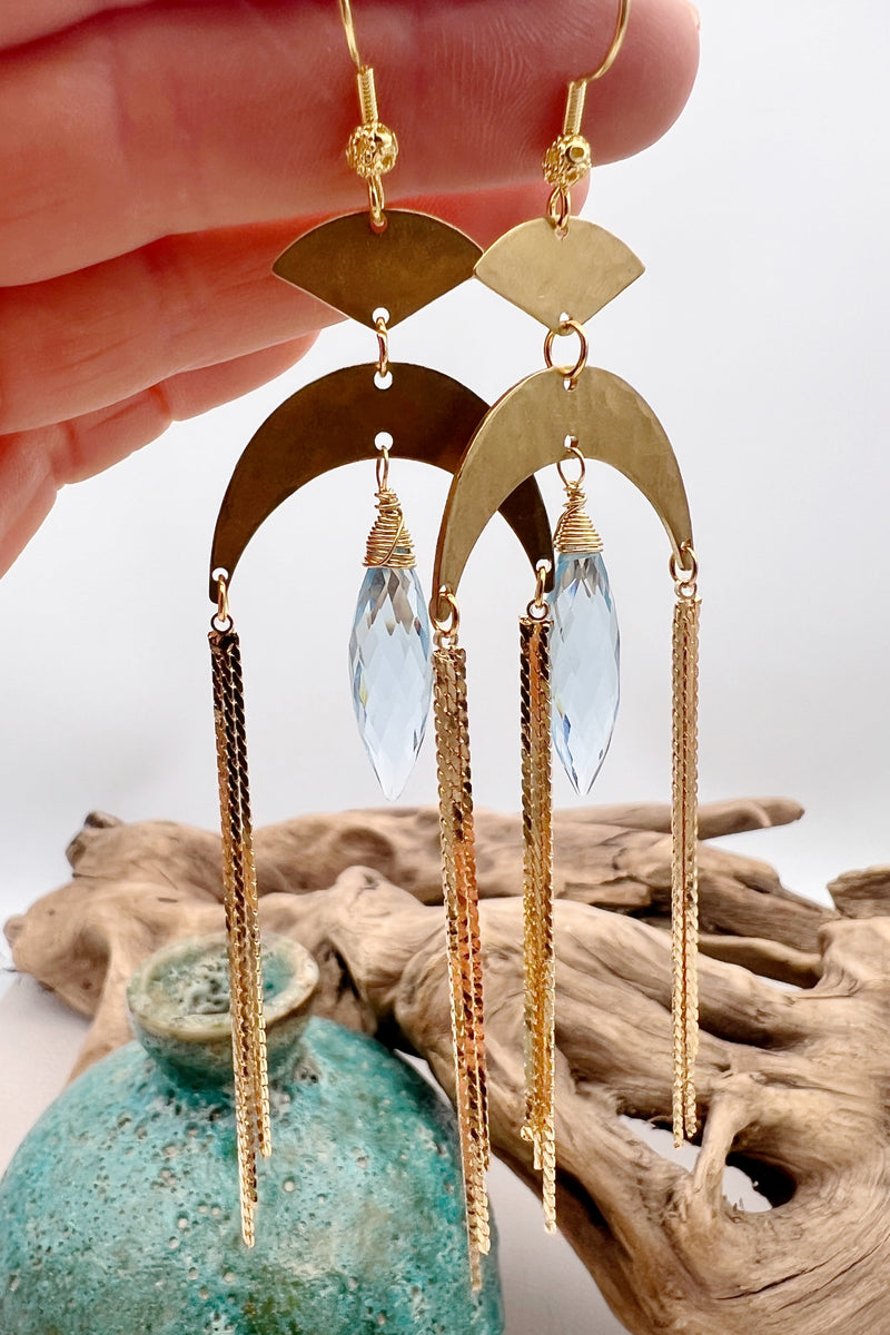 Gold statement earrings with a faceted aqua crystal, a celestial moon charm, and flowing golden tassels. Bold, lightweight, and designed for movement and shine.