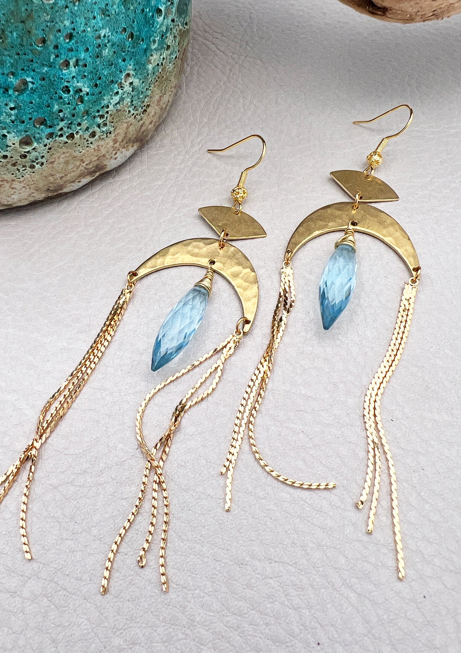 Gold statement earrings with a faceted aqua crystal, a celestial moon charm, and flowing golden tassels. Bold, lightweight, and designed for movement and shine.