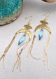 Gold statement earrings with a faceted aqua crystal, a celestial moon charm, and flowing golden tassels. Bold, lightweight, and designed for movement and shine.