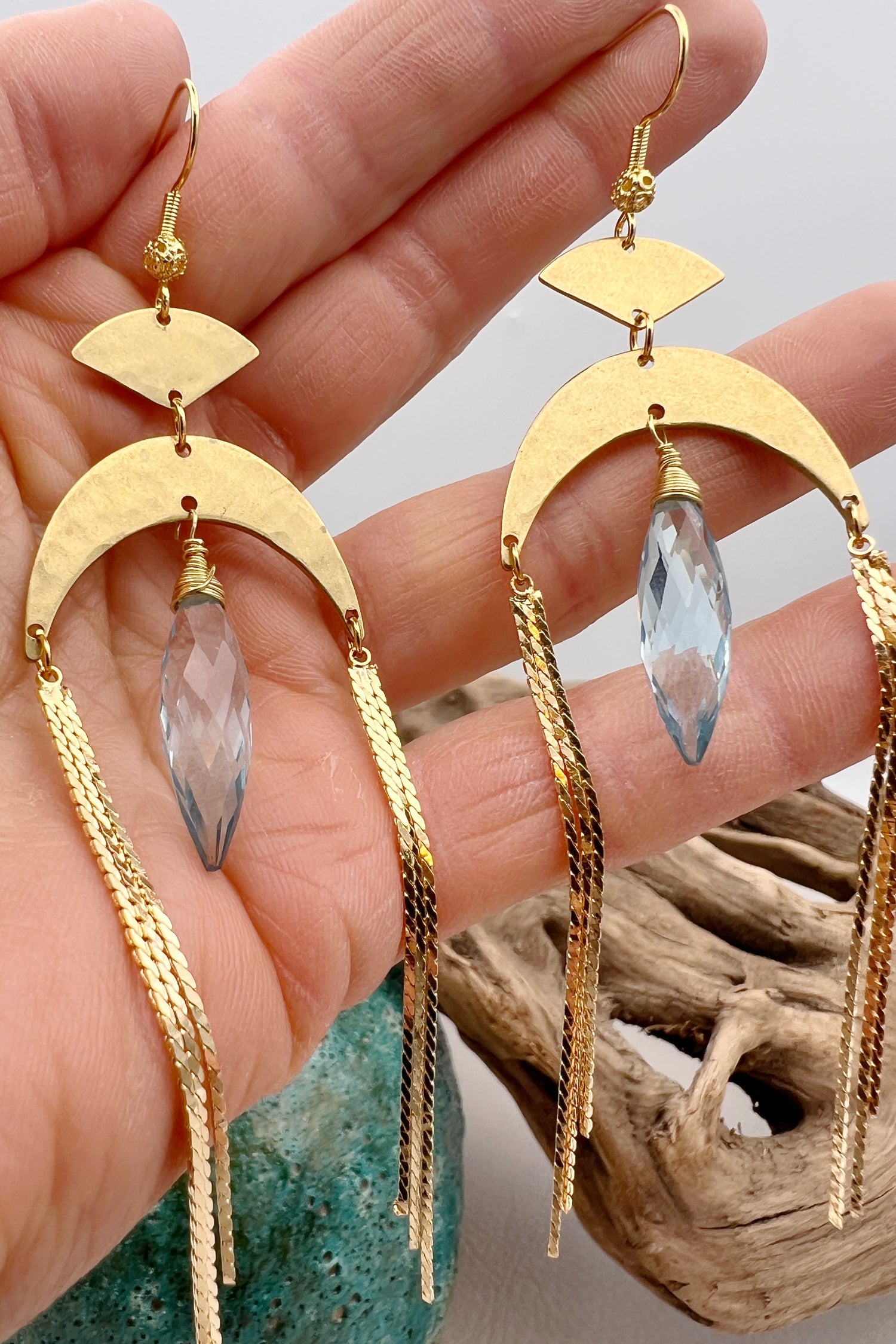 Gold statement earrings with a faceted aqua crystal, a celestial moon charm, and flowing golden tassels. Bold, lightweight, and designed for movement and shine.