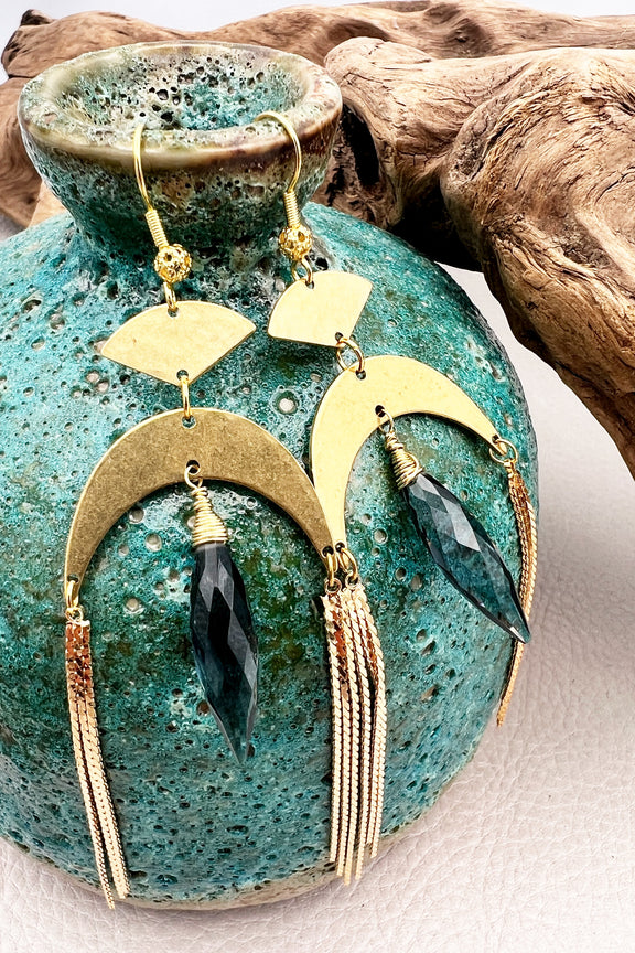 Gold statement earrings with a faceted aqua crystal, a celestial moon charm, and flowing golden tassels. Bold, lightweight, and designed for movement and shine.