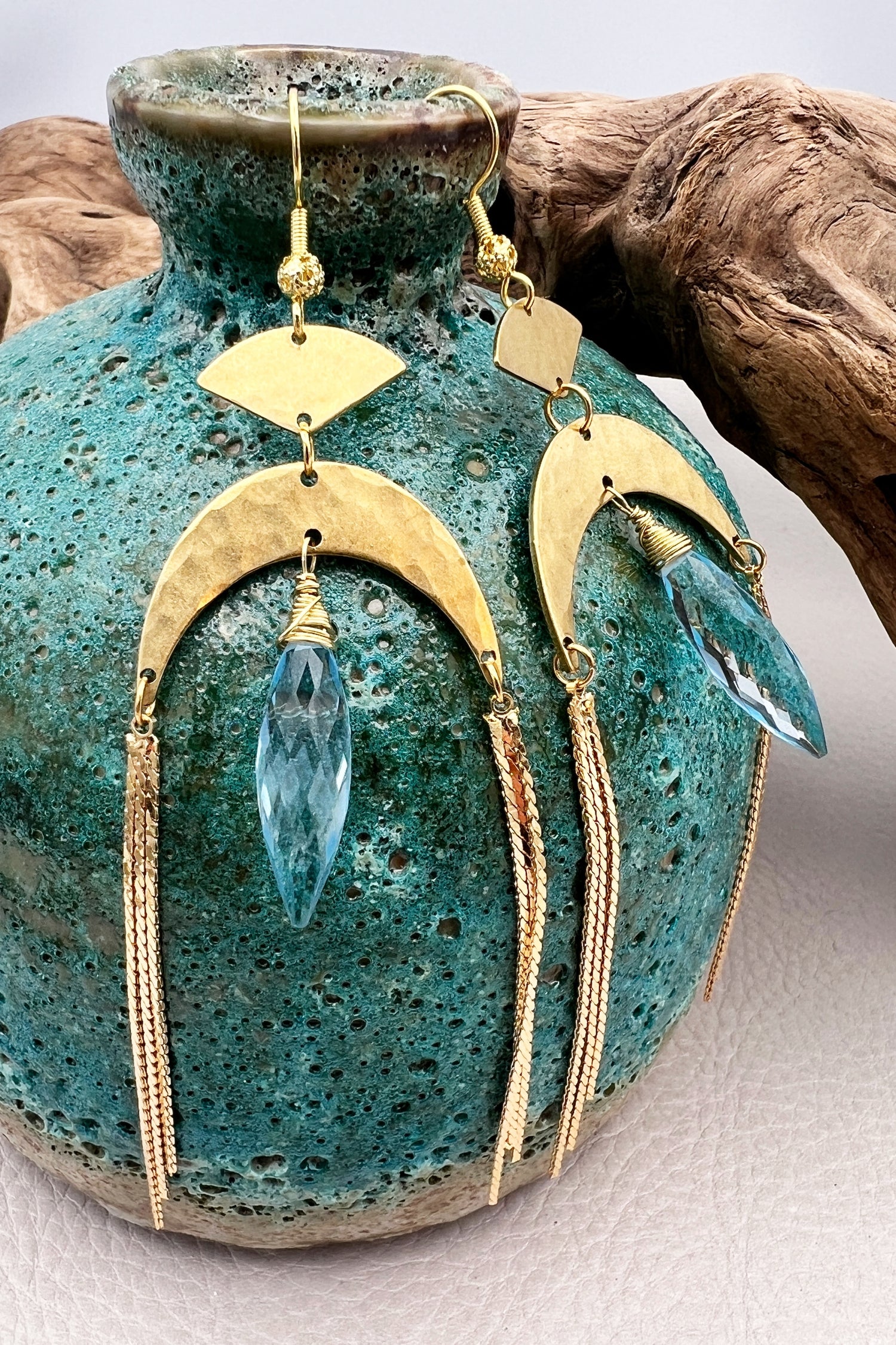 Gold statement earrings with a faceted aqua crystal, a celestial moon charm, and flowing golden tassels. Bold, lightweight, and designed for movement and shine.